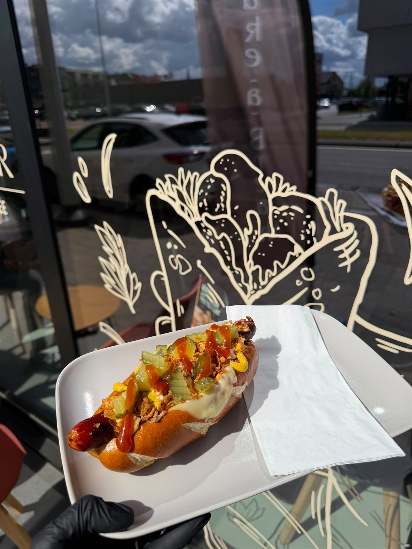 Hotdog met augurk, gefruite ui en sauzen op een bord voor het raam van de zaak